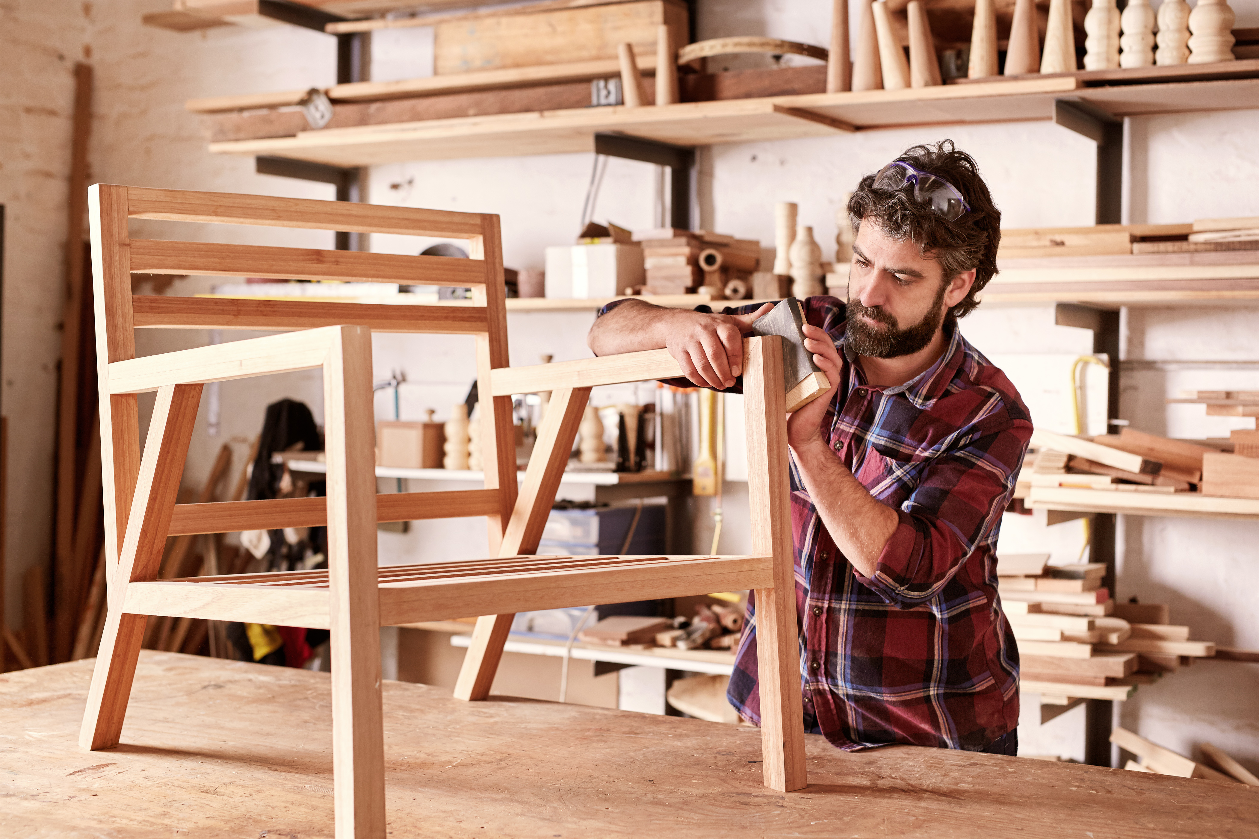 Serious furniture designer carefully sanding a chair frame that he is busy manufacturing in his woodwork studio, with shelves of wooden items behind him