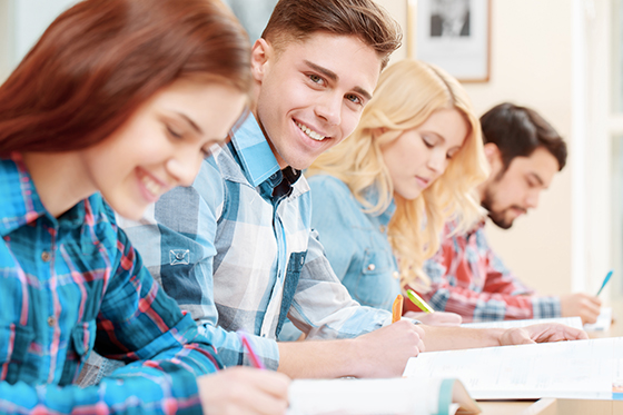 Got the answer. Young handsome student in selective focus smiling at a camera while sitting at the desk with his classmates